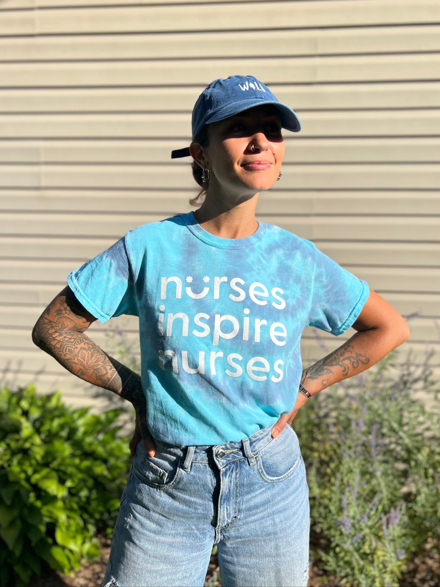 Person wearing a blue 'nurses inspire nurses' t-shirt and cap outdoors.